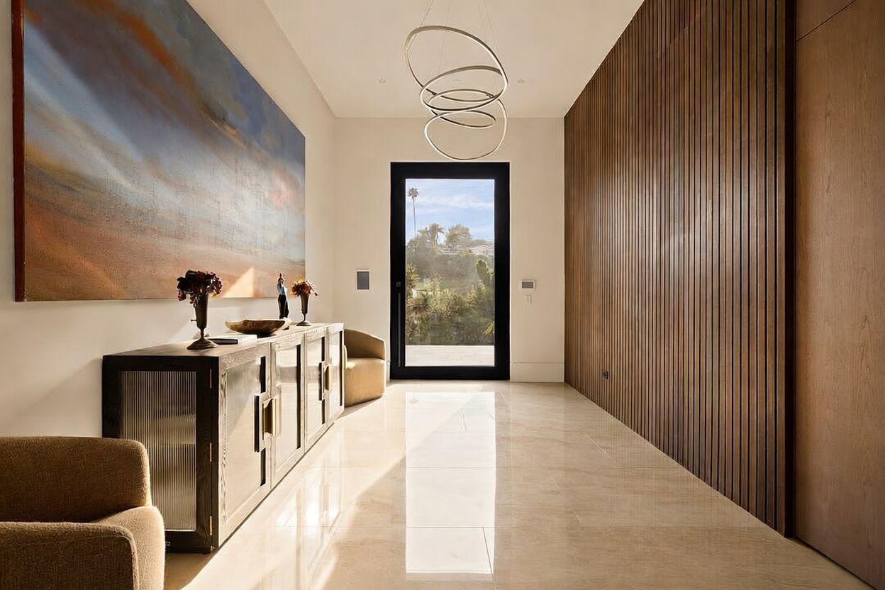 This sophisticated entryway features a polished stone floor and a striking wall of vertical wood slats that adds texture and warmth to the space. A large, abstract landscape painting hangs above a dark console table, while a modern, sculptural light fixture hangs from the ceiling. The perspective looks directly toward a large glass-paneled front door that provides a clear view of the outdoor landscape.