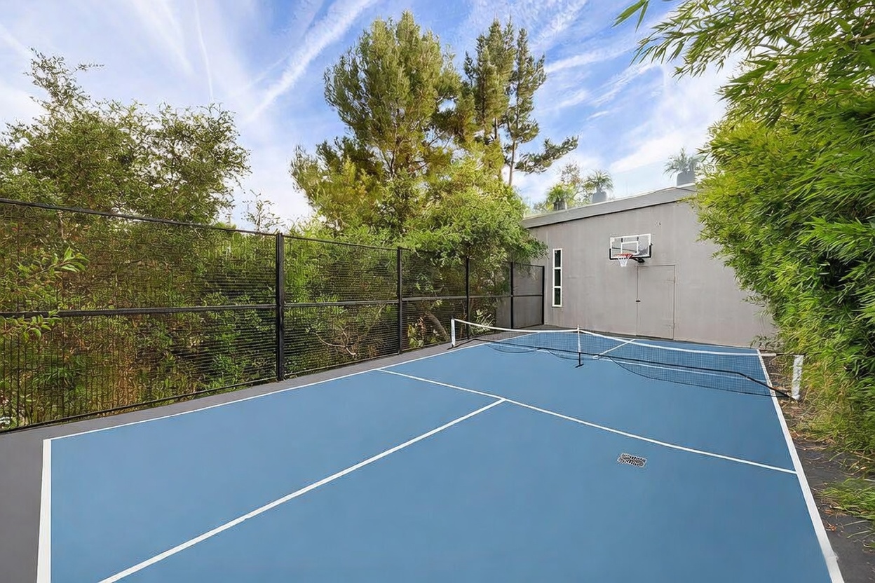 This outdoor space features a multi-purpose sports court with a blue surface, a tennis net, and a basketball hoop mounted on a gray wall. The court is enclosed by a black metal fence and surrounded by lush green trees and bamboo, creating a private recreational area. The perspective is from the court level, capturing the functional layout and the integration of sports amenities within a landscaped setting.