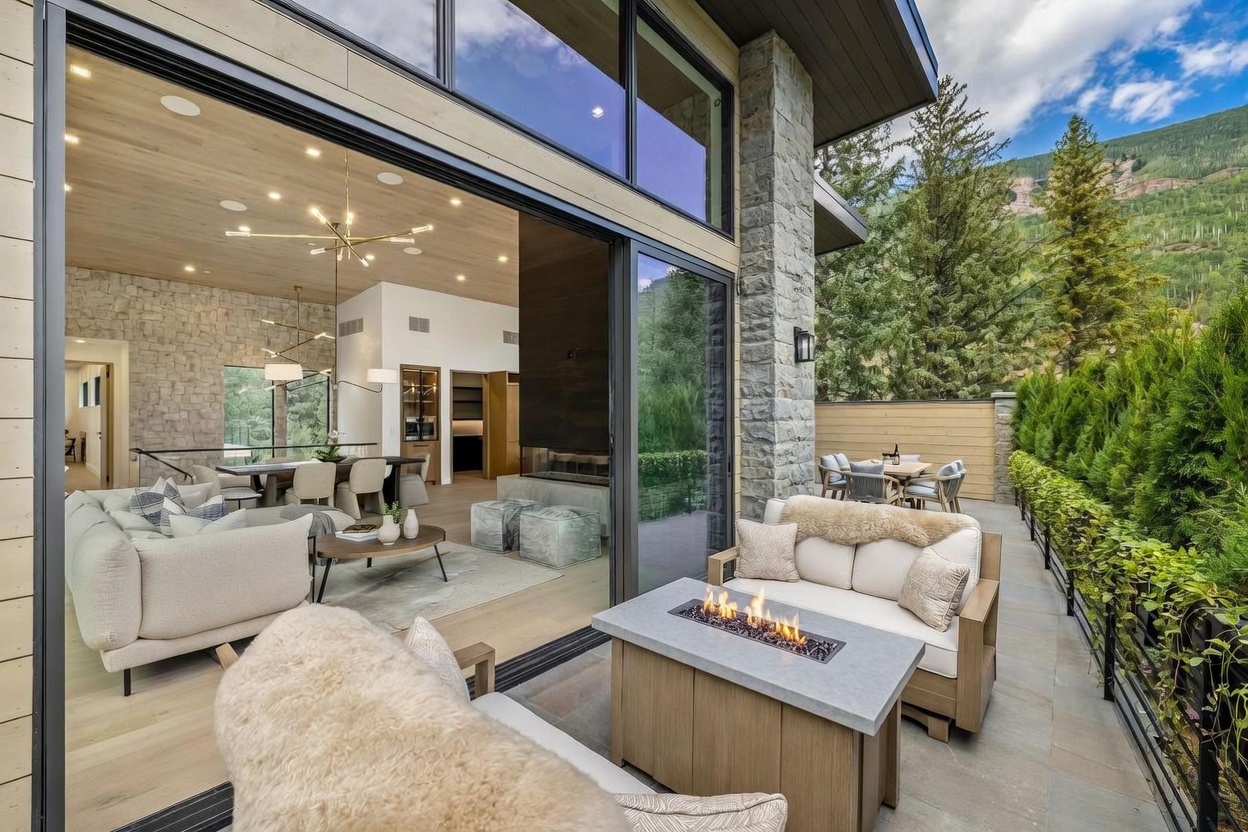 This image captures a seamless transition between a modern indoor living area and an outdoor patio space, facilitated by large sliding glass doors. The patio features a cozy seating arrangement with a fire pit, while the interior showcases a contemporary living room with light wood ceilings and stone-clad walls. The setting is framed by lush greenery and mountain views, creating a sophisticated and inviting atmosphere for relaxation.