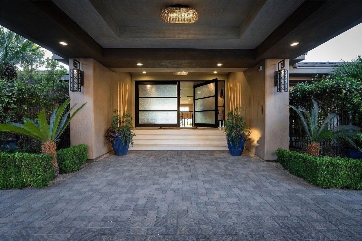 This grand entryway features a covered porch with a textured stone-paver driveway leading to a set of modern, multi-pane glass double doors. The entrance is framed by symmetrical pillars adorned with decorative wall-mounted lanterns and large potted plants with bamboo accents. The perspective is centered and eye-level, highlighting the sophisticated, welcoming architecture of the home's facade.