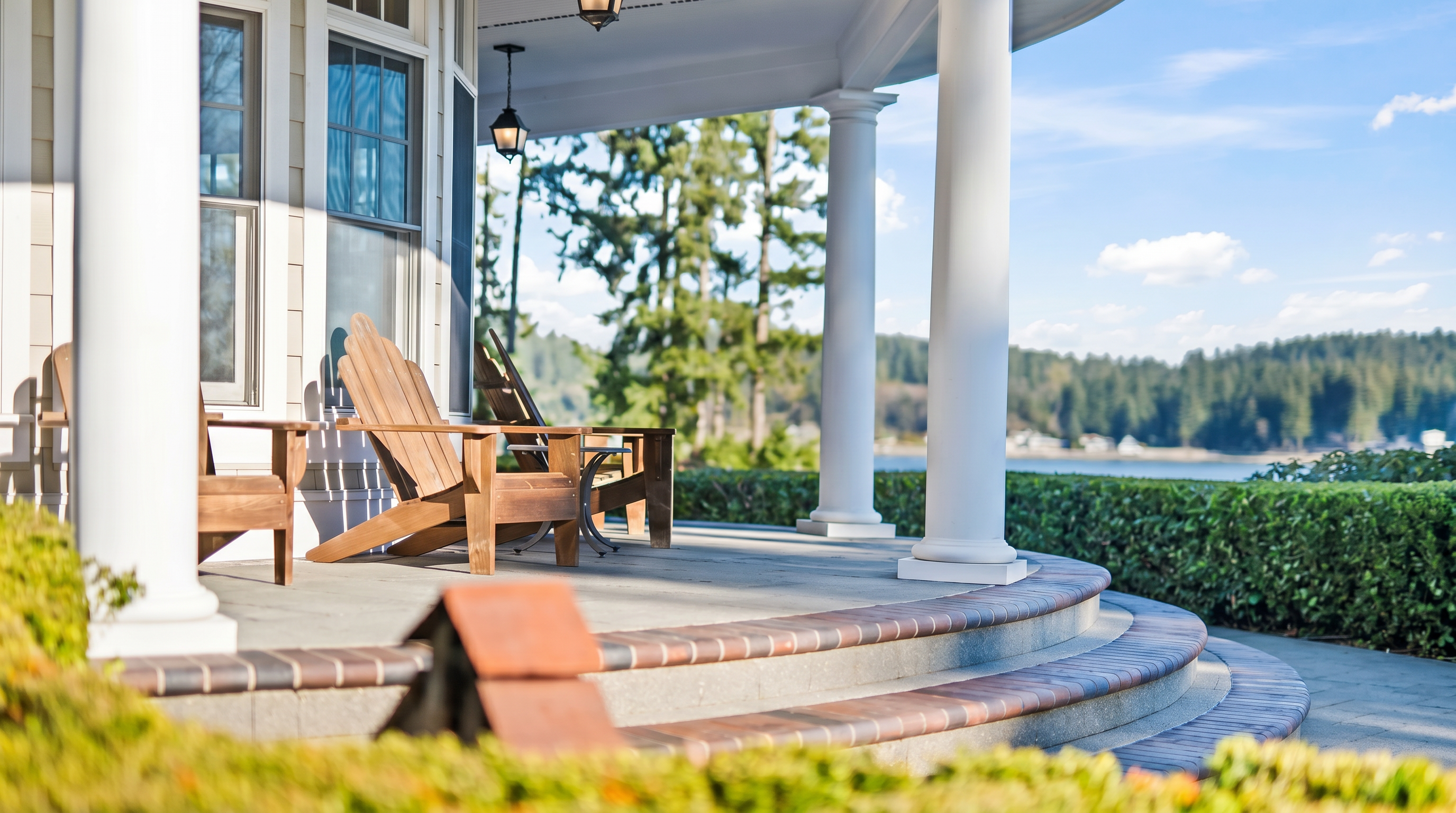 This inviting covered porch features classic white columns and a curved brick-edged stone floor, creating a sophisticated outdoor living space. Two wooden Adirondack chairs are positioned to overlook a lush green hedge and a serene waterfront view in the distance. The perspective is low and cinematic, emphasizing the architectural details of the porch and the peaceful, scenic surroundings.