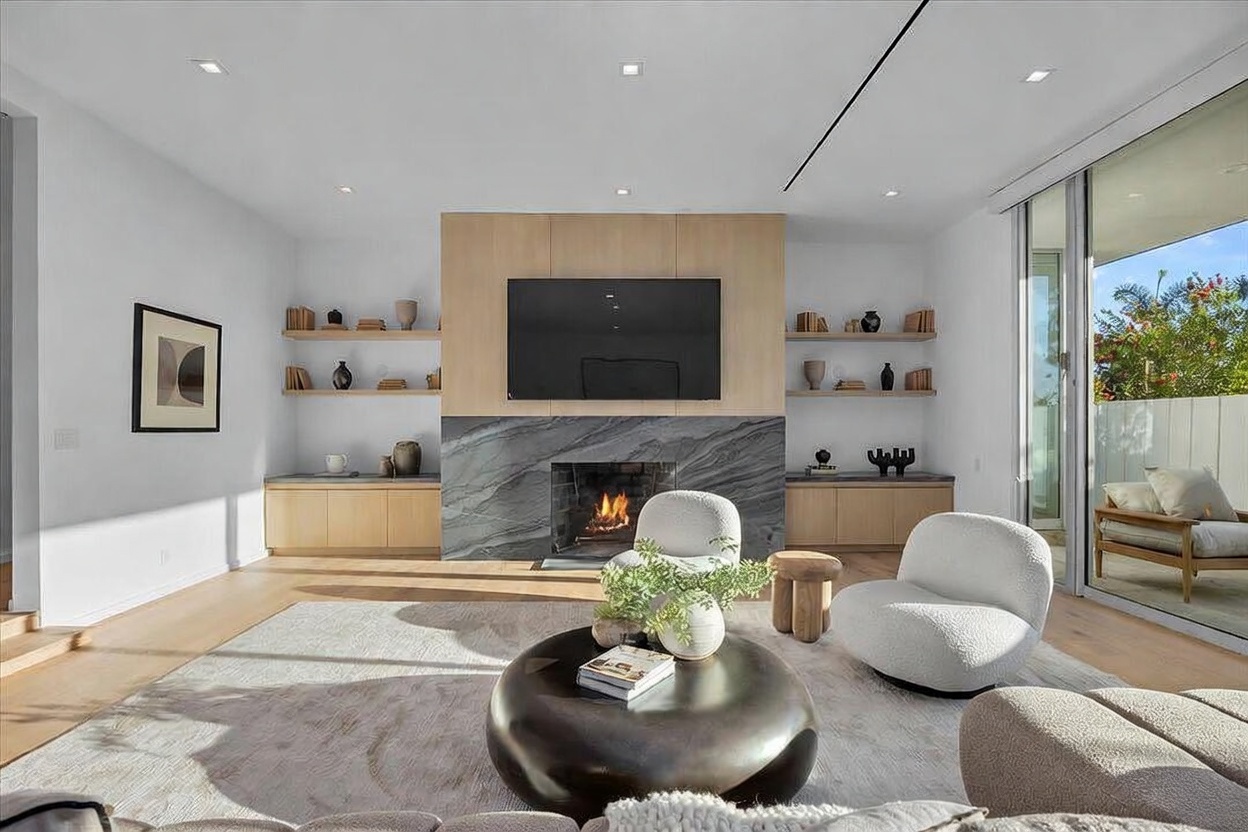 This bright and airy living room features a central fireplace with a striking, dark-veined stone surround that extends upward to meet the ceiling. The space is anchored by a large, dark circular coffee table and two textured, cream-colored lounge chairs, all set upon a neutral area rug. Light wood cabinetry flanks the fireplace, providing integrated storage and open shelving for decor, while large glass sliding doors to the right invite natural light and provide a view of the outdoor patio.
