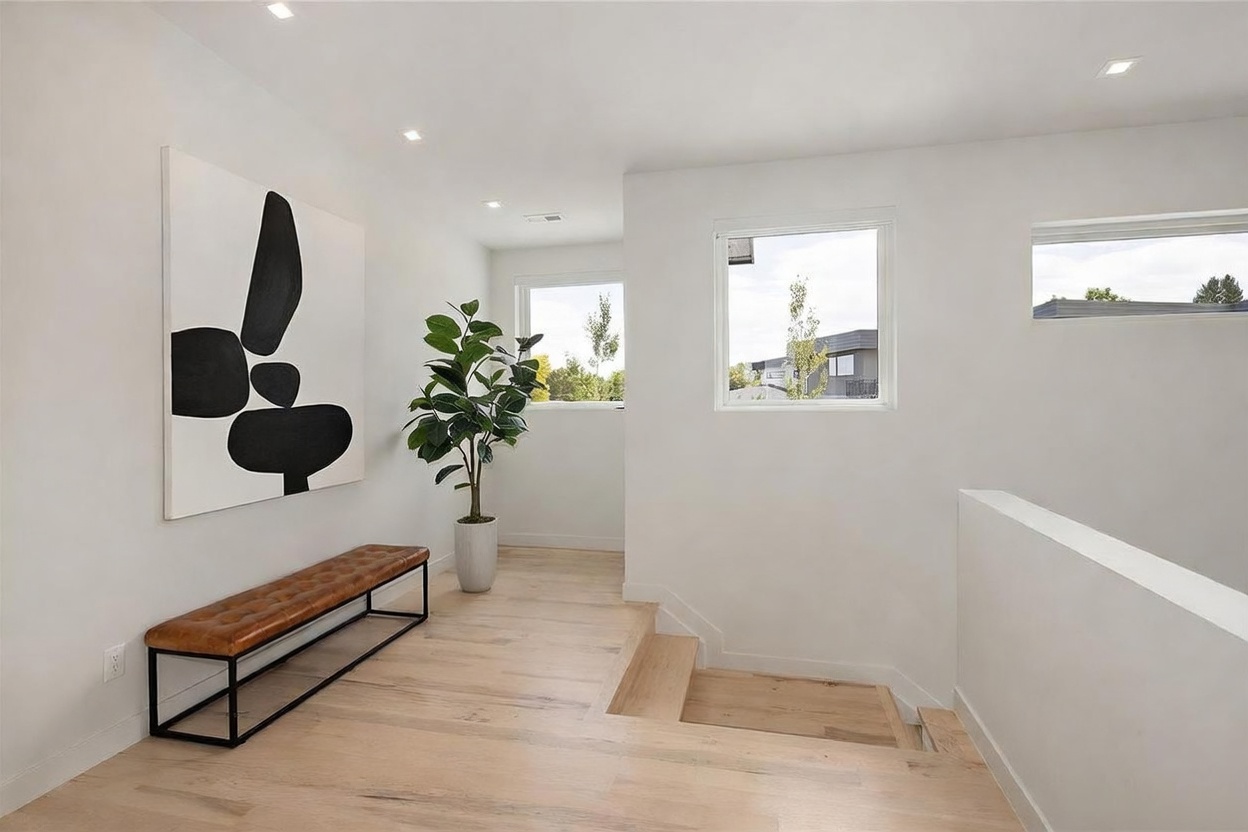 This bright, modern landing area features light wood flooring and crisp white walls, creating an airy and minimalist aesthetic. A leather-upholstered bench sits beneath a large, abstract black-and-white art piece, while a potted plant adds a touch of greenery near the staircase. The space is illuminated by recessed lighting and large windows that provide natural light and views of the surrounding neighborhood.
