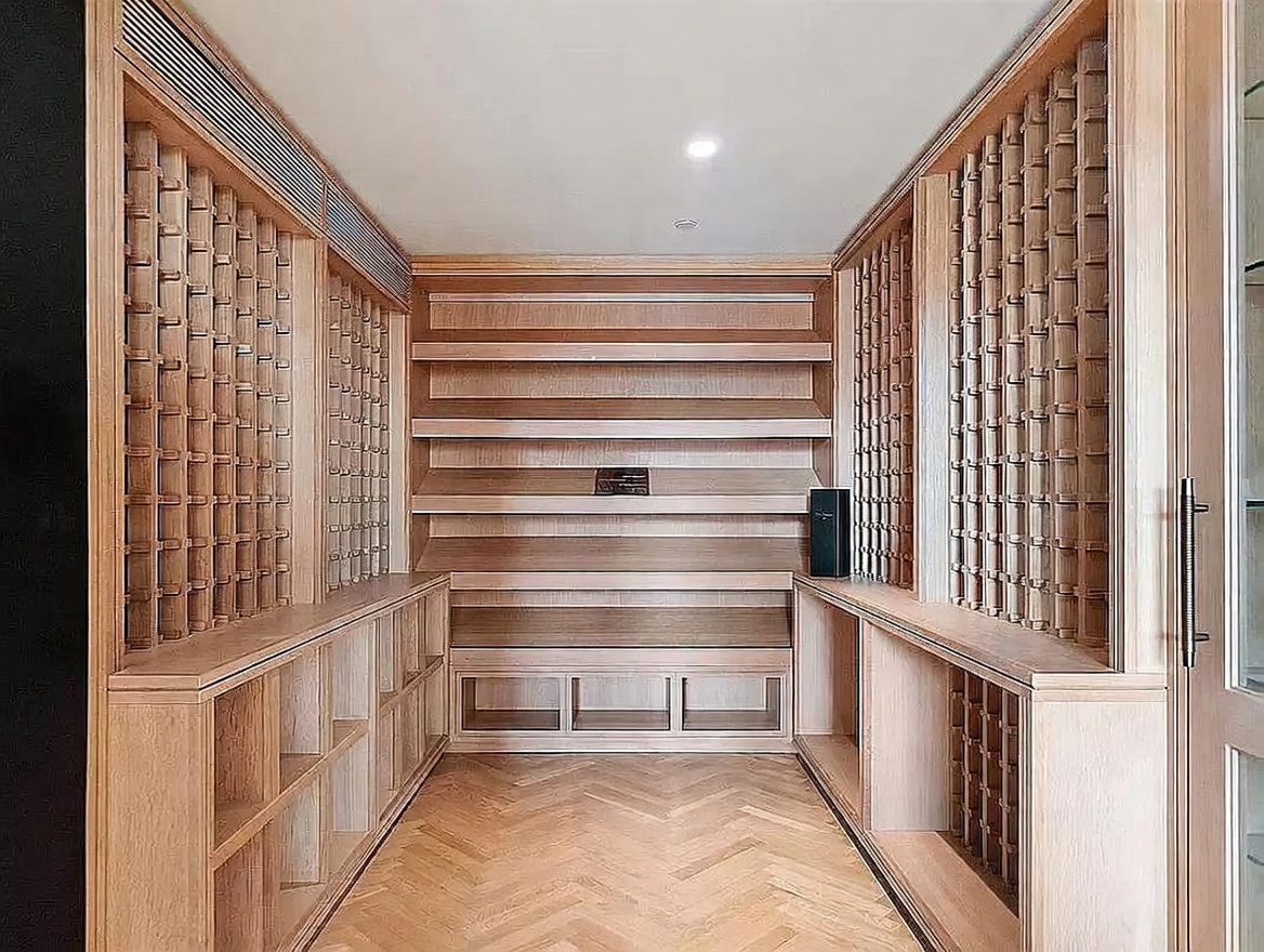 This sophisticated wine cellar features extensive custom light wood cabinetry designed for bottle storage, including both individual racks and open shelving. The space is finished with a classic herringbone-patterned wood floor and recessed lighting, creating a warm and organized atmosphere. The symmetrical layout and clean lines provide a refined, high-end aesthetic suitable for a dedicated collection.