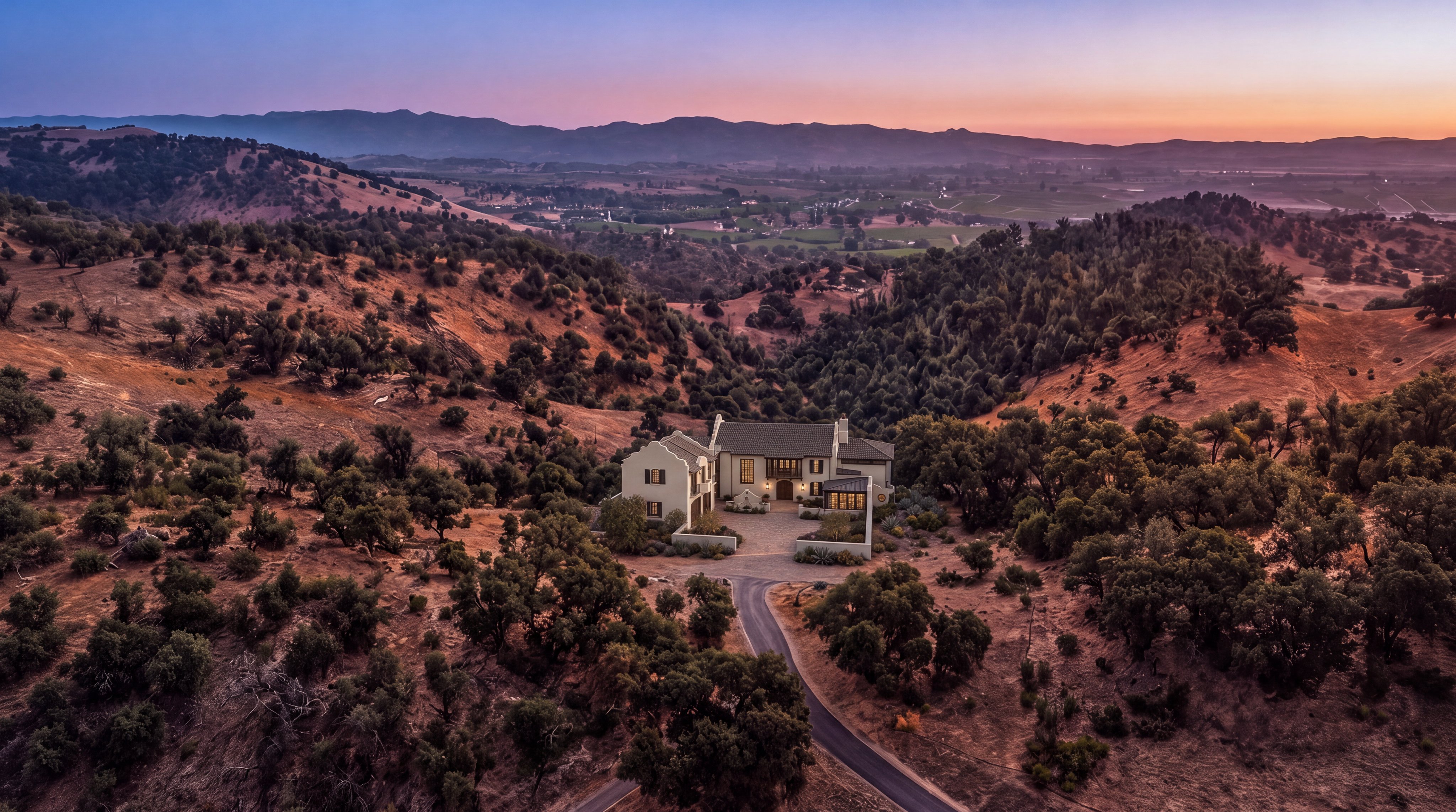 This stunning aerial view captures a luxury villa nestled within a secluded, rolling landscape of golden hills and mature trees. The home features a distinct Mediterranean-inspired architectural style with a paved courtyard, multi-gabled roofline, and warm, illuminated interiors that create an inviting atmosphere. The elevated perspective highlights the property's expansive, private setting and picturesque views of the distant valley at dusk.