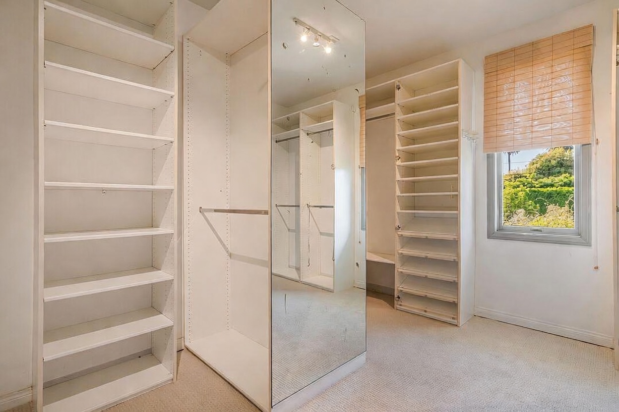 This organized walk-in closet features extensive white shelving and hanging rods, providing ample storage space. A large, full-length mirror is integrated into the center cabinetry, reflecting the opposite side of the room and adding a sense of depth. A window with a bamboo shade allows natural light to brighten the space, which is finished with neutral carpeting.