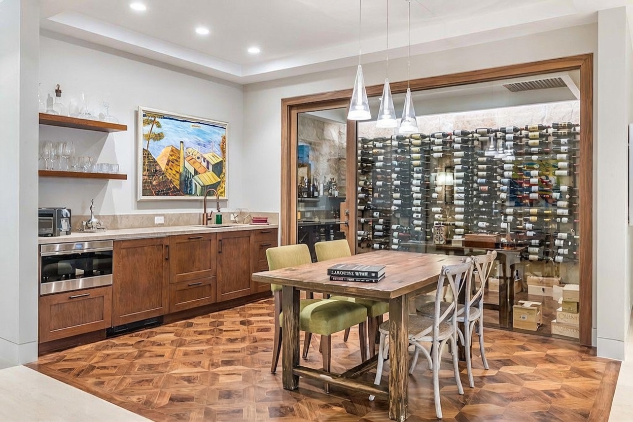 This sophisticated wine tasting room features a custom glass-enclosed wine cellar as its focal point, showcasing an extensive collection of bottles. A rustic wooden dining table sits in the foreground, complemented by patterned wood flooring and a wet bar area with open shelving and cabinetry. The space is illuminated by modern pendant lighting, creating an elegant and inviting atmosphere for entertaining.