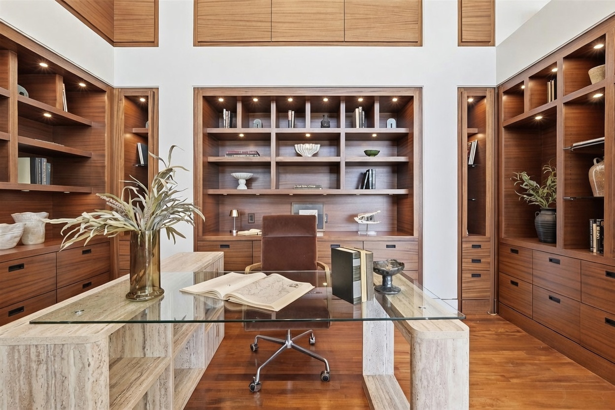 This sophisticated home office features extensive custom wood cabinetry and shelving that spans the entire wall, providing ample storage and display space. A central glass-topped desk with stone-textured supports sits in the foreground, complemented by a brown leather office chair. The room is bathed in warm, recessed lighting, creating a professional yet inviting atmosphere suitable for a high-end residence.