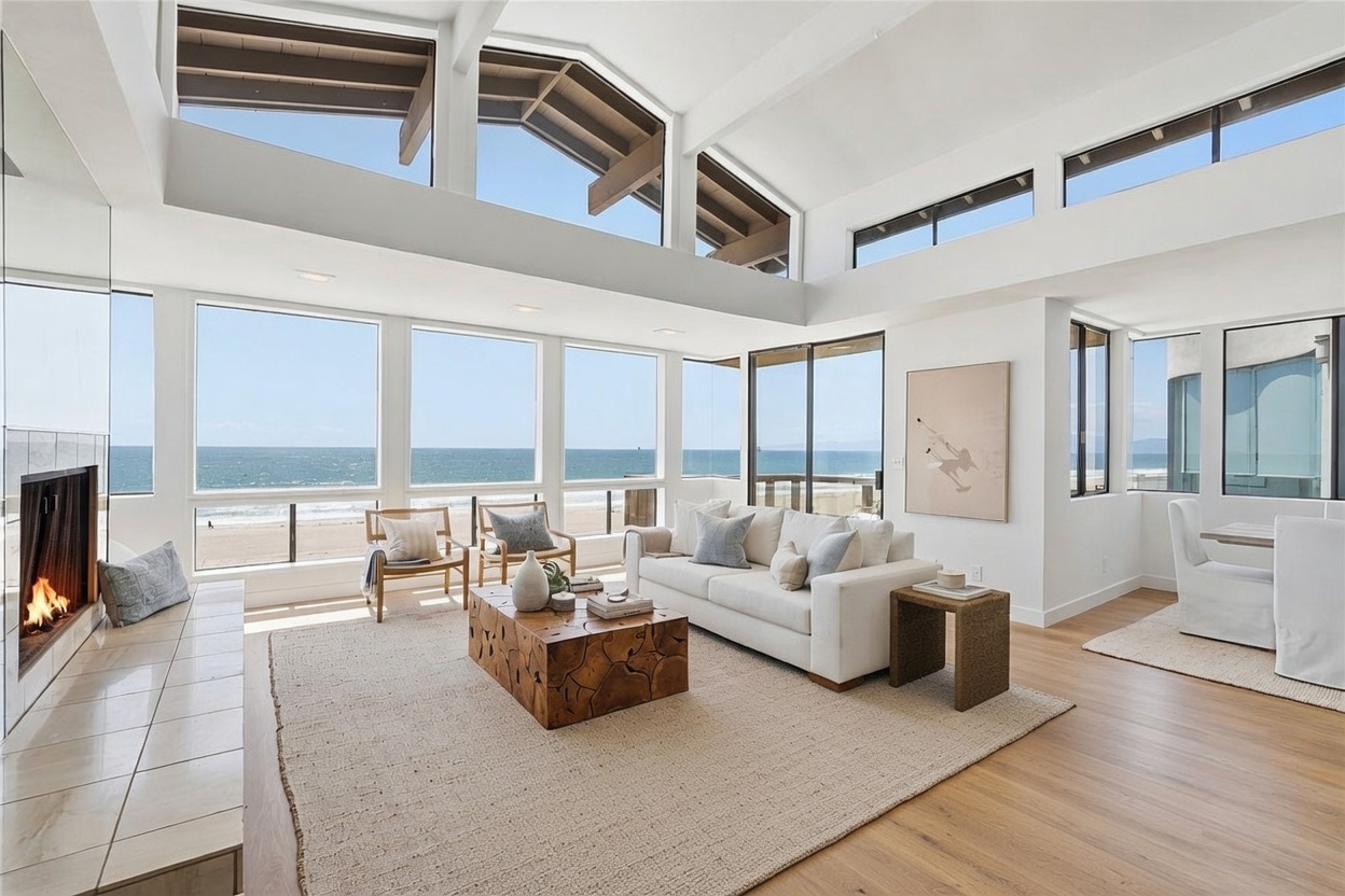 This bright and airy living room features a neutral color palette and a large, natural wood coffee table as a central focal point. The space is defined by expansive windows that offer panoramic ocean views, complemented by a tall fireplace surround on the left. The open-concept layout includes comfortable seating arrangements and light wood flooring, creating a serene and sophisticated coastal atmosphere.
