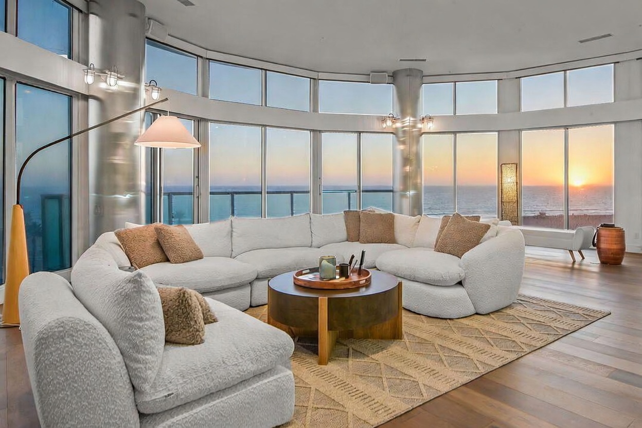 This spacious living room features a large, curved white sectional sofa arranged around a circular wooden coffee table on a textured area rug. The room is defined by expansive floor-to-ceiling windows that offer a panoramic view of the ocean at sunset, bathing the space in warm, natural light. The interior design is contemporary and airy, complemented by light wood flooring and minimalist structural columns.