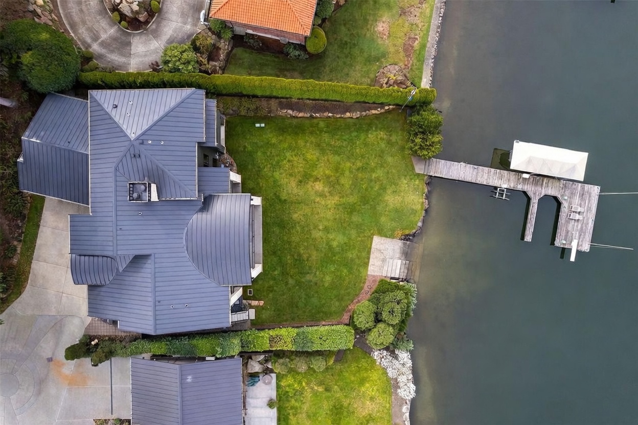 This high-angle aerial perspective captures a waterfront property featuring a large home with a dark metal roof and a well-manicured lawn. A private wooden dock extends from the grassy backyard into the calm water, complete with a covered boat slip. The property is bordered by lush green hedges, providing a sense of privacy and a clear transition between the landscaped grounds and the lake.