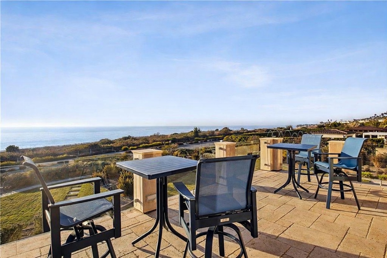 This elevated outdoor patio features stone pavers and multiple sets of bistro-style tables and chairs, perfectly positioned to take advantage of the expansive ocean views. The space is bordered by a glass railing that maintains an unobstructed sightline toward the horizon and the coastal landscape below. The bright, sunny atmosphere highlights the serene and luxurious nature of this coastal living space.