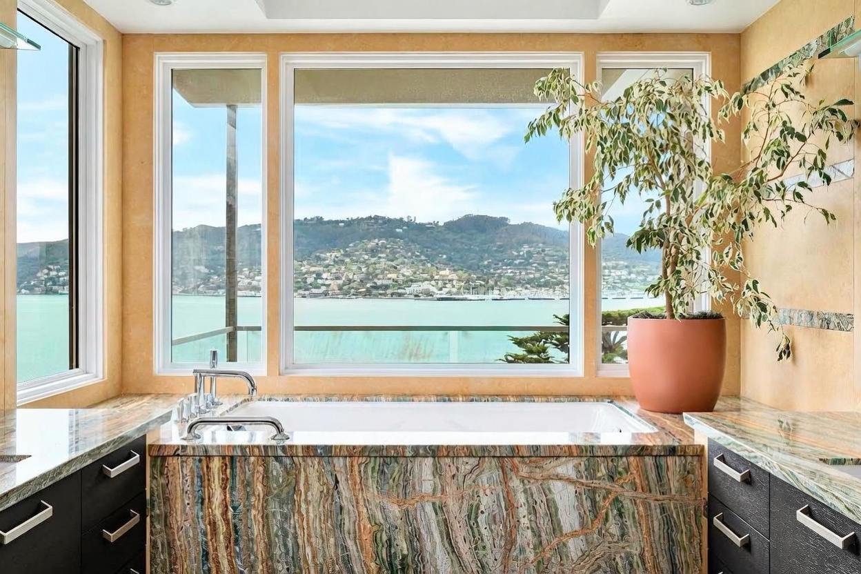 This luxurious primary bathroom features a prominent soaking tub encased in striking, multi-colored patterned stone that serves as the room's focal point. Large windows frame a panoramic view of the bay and distant hills, bathing the space in natural light. Dark cabinetry with sleek hardware flanks the tub, creating a sophisticated contrast against the warm, textured wall finishes.