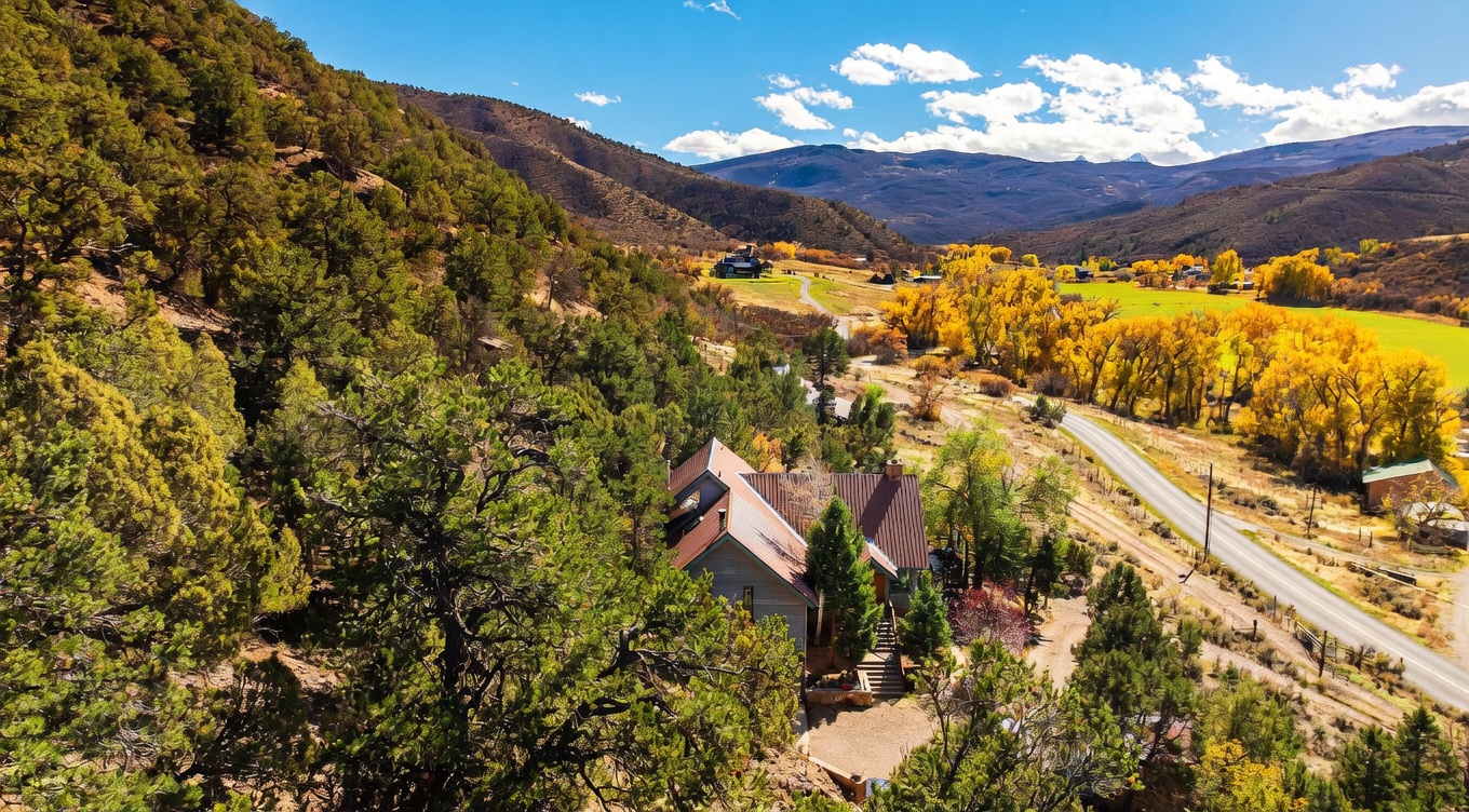 This high-angle aerial view captures a rustic home nestled into a hillside covered with dense, mature trees. The property features a multi-gabled roofline and a wooden deck, overlooking a scenic valley with a winding road, vibrant autumn foliage, and distant mountain peaks. The perspective emphasizes the home's secluded, natural setting and its integration into the surrounding landscape.