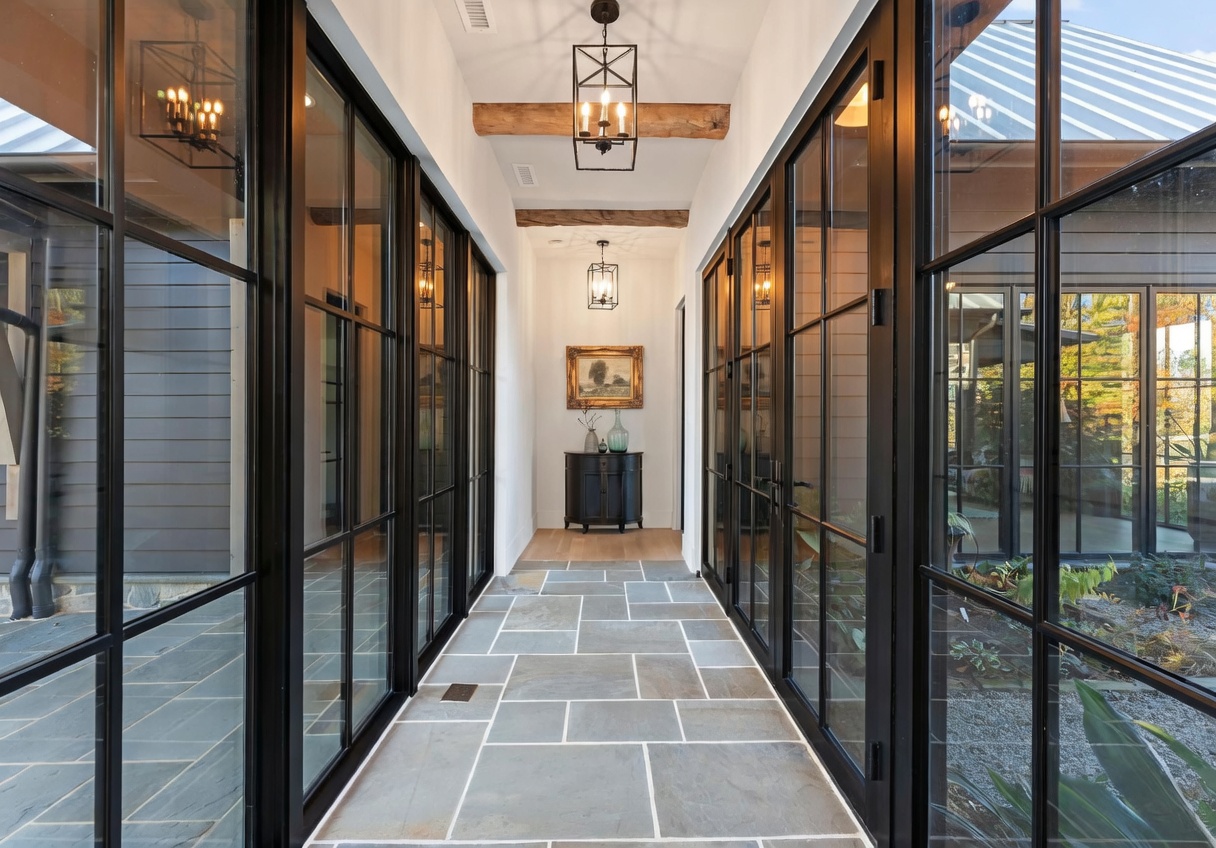 This elegant, light-filled hallway features floor-to-ceiling glass walls on both sides, offering a seamless view of the surrounding outdoor landscape. The space is defined by a stone-tiled floor, exposed wooden ceiling beams, and two classic lantern-style pendant lights. At the end of the corridor, a dark console table topped with decorative vases and a framed painting creates a sophisticated focal point.