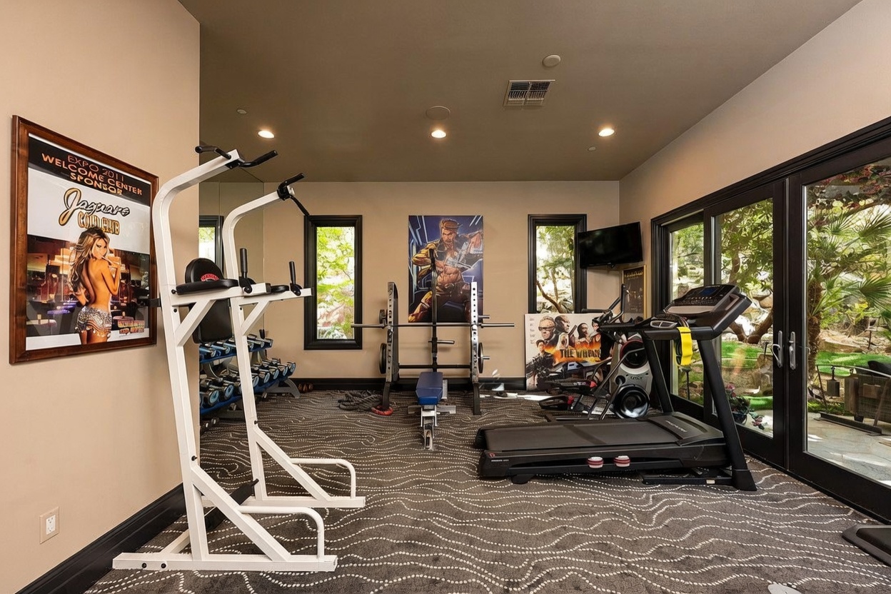 This home gym features a patterned carpet and is equipped with a white pull-up and dip station, a weight bench with a barbell, and a treadmill positioned near large glass sliding doors. The room is decorated with framed posters on the walls and recessed lighting, creating a functional workout space with a view of the outdoor greenery. The perspective is a wide shot from the corner of the room, capturing the layout of the exercise equipment.