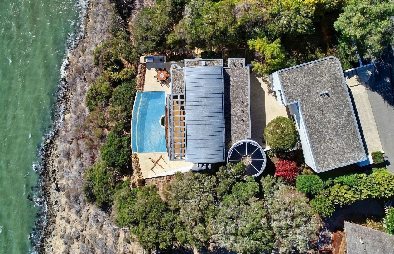 This high-angle aerial perspective captures a unique modern residence perched on a coastal cliffside overlooking the water. The property features a distinctive curved metal roof, a private swimming pool with a deck area, and a circular architectural element integrated into the surrounding lush landscaping. The layout emphasizes a seamless connection between the home's geometric design and the rugged natural terrain of the shoreline.