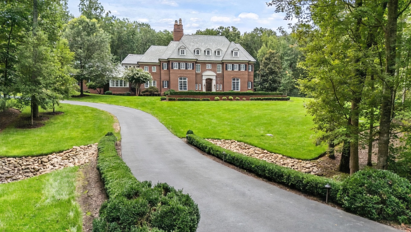This stately brick colonial-style residence is set back from a winding paved driveway, surrounded by lush green lawns and mature trees. The home features a symmetrical facade with multiple dormer windows, a prominent central chimney, and a manicured hedge lining the front garden bed. The perspective is a wide-angle shot from the driveway, emphasizing the grand scale and private, wooded setting of the property.