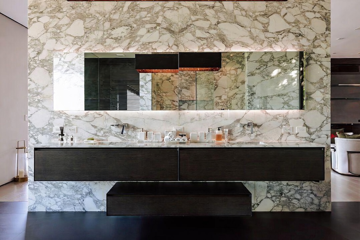 This luxurious primary bathroom features a striking floor-to-ceiling marble wall that serves as a dramatic backdrop for a sleek, dark wood double vanity. The space is illuminated by integrated mirror lighting and a modern, textured pendant fixture, creating a sophisticated and high-end atmosphere. The clean lines and minimalist design emphasize a contemporary aesthetic, perfect for a high-end residential property.
