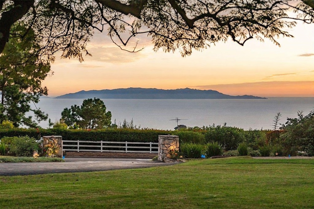 This scenic outdoor view captures a lush green lawn leading toward a white fence and stone-pillared gate, framed by the overhanging branches of a large tree. In the distance, a serene ocean horizon is dominated by a prominent island silhouette under a soft, golden-hued sunset sky. The perspective is from a slightly elevated position, emphasizing the expansive and tranquil nature of the property's landscape.