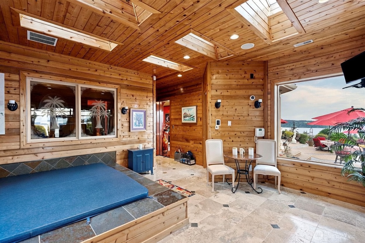 This sun-drenched indoor spa room features extensive wood paneling on the walls and ceiling, complemented by multiple skylights that flood the space with natural light. A large, tiled hot tub platform sits prominently in the foreground, while a small bistro set provides a cozy seating area near a large window overlooking a scenic outdoor patio and water view. The room combines rustic wood textures with light-toned tile flooring, creating a relaxing and inviting atmosphere.