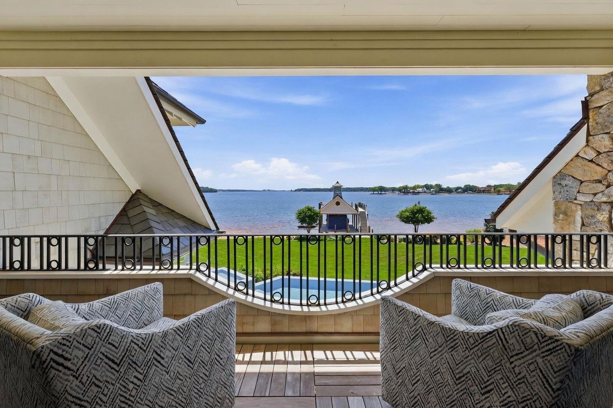 This elevated balcony offers a serene, framed view of a calm lake and a private boat dock. The space is furnished with two comfortable, patterned armchairs and features a decorative metal railing that curves inward to enhance the sightline. The setting provides a peaceful, high-end outdoor living experience overlooking a lush green lawn and the water beyond.