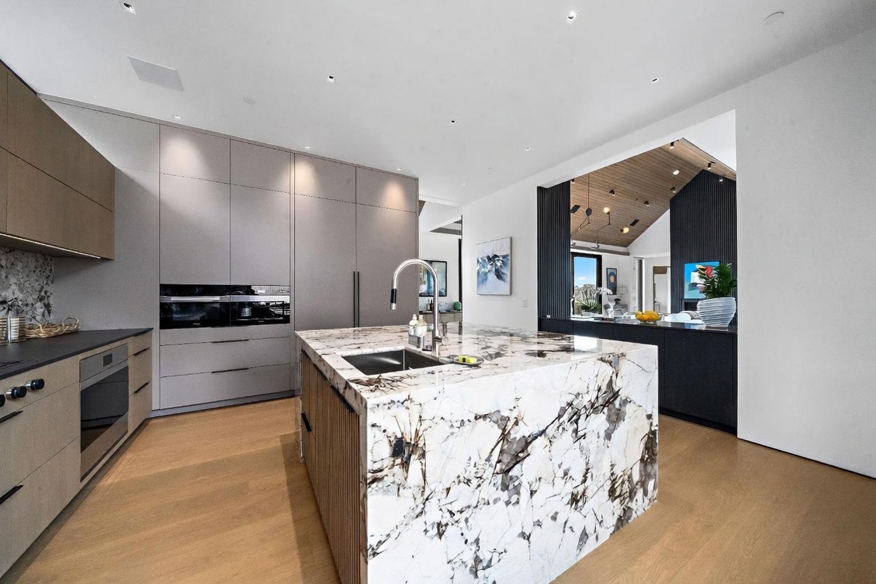 This modern kitchen features a large central island with a striking patterned stone countertop and a built-in sink. The space is equipped with sleek, flat-panel cabinetry in a neutral tone, integrated appliances, and light wood flooring. An open transition leads into an adjacent living area with a vaulted ceiling, creating an airy and sophisticated open-concept layout.