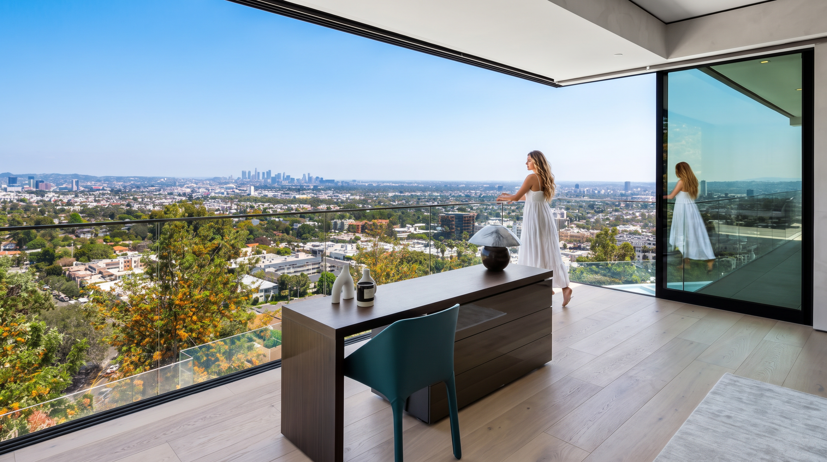 This modern home office offers a breathtaking, unobstructed view of the Los Angeles skyline through floor-to-ceiling glass walls that lead to an expansive balcony. The space features a sleek, dark wood executive desk, a contemporary teal accent chair, and minimalist decor, creating a sophisticated and inspiring work environment. The seamless integration of indoor and outdoor living spaces emphasizes the property's luxury status and prime location.