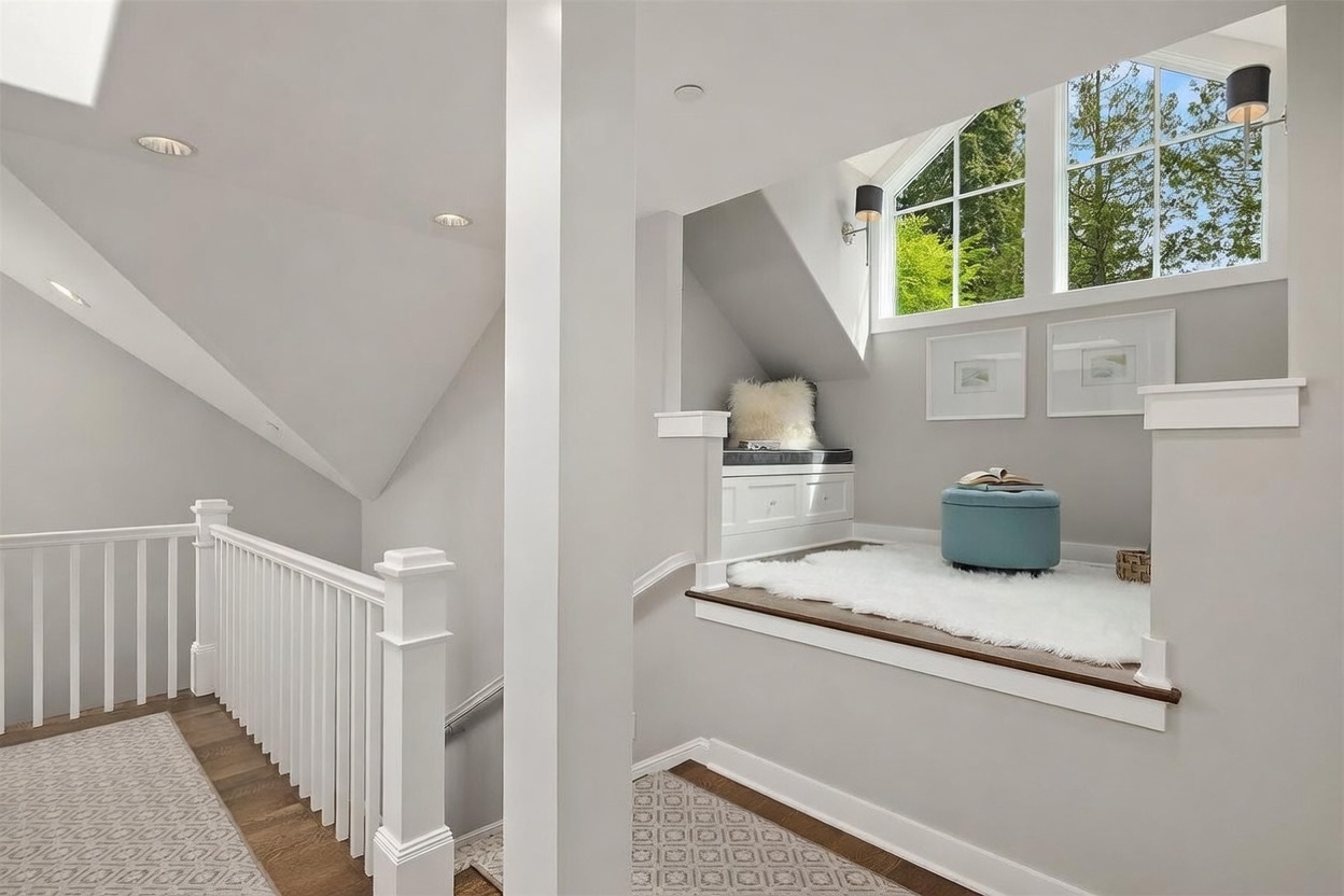 This bright and airy landing area features a cozy built-in reading nook tucked under a sloped ceiling, complete with a plush white rug and a soft blue ottoman. A white wooden staircase railing leads down to the lower level, while large windows above the nook flood the space with natural light. The neutral color palette and clean lines create a serene and inviting atmosphere, perfect for a quiet reading corner.