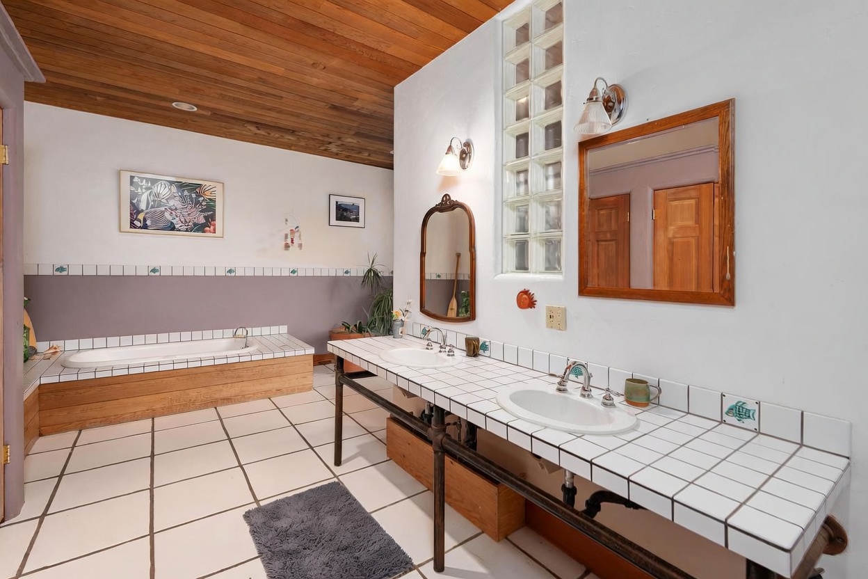 This spacious bathroom features a dual-sink vanity with a tiled countertop and an exposed pipe base, paired with a separate built-in soaking tub. The room is accented by a vertical glass block window, a wood-paneled ceiling, and decorative fish-themed tiles. The perspective captures the entire vanity area and the tub, showcasing a unique, rustic-inspired design.