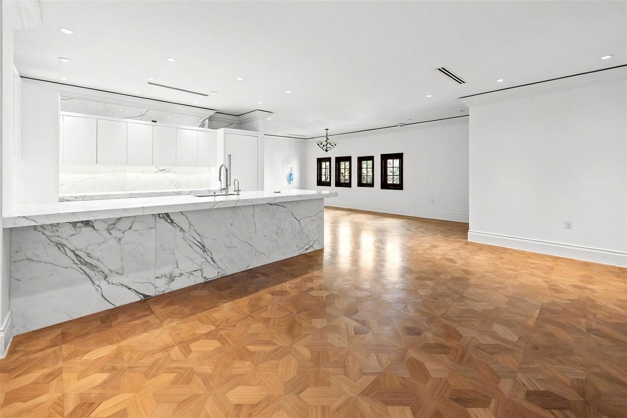 This bright, open-concept kitchen features a large island with a prominent stone waterfall edge and matching backsplash. The space is characterized by clean white cabinetry, recessed lighting, and intricate patterned wood flooring. A series of dark-framed windows lines the far wall, providing a sharp contrast to the minimalist white walls and creating a spacious, airy atmosphere.