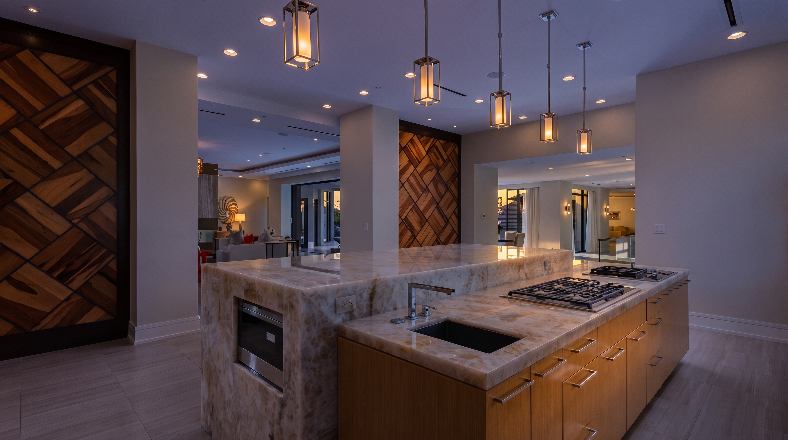 This modern kitchen features a large central island with a stone countertop, an integrated sink, and a built-in gas cooktop. The space is accented by light wood cabinetry and decorative wood paneling on the walls, illuminated by three hanging pendant lights. The open-concept design provides a clear view into the adjacent living area, creating a bright and airy atmosphere.