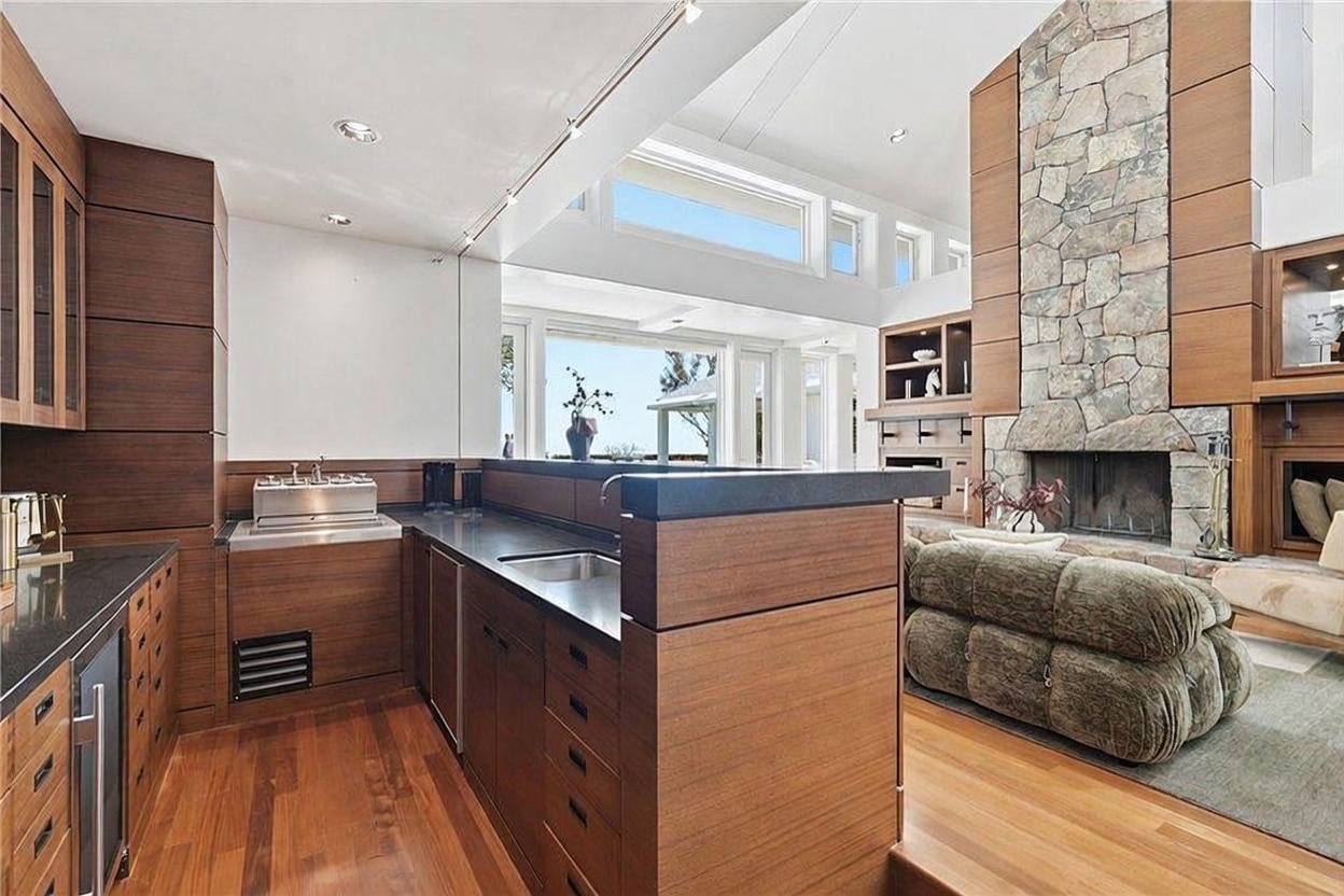 This open-concept kitchen features warm-toned wood cabinetry, dark countertops, and a built-in stainless steel grill station. The space flows directly into a living area anchored by a prominent stone-clad chimney breast, creating a cohesive and inviting atmosphere. Large windows and clerestory glass allow for abundant natural light, highlighting the rich wood flooring and the seamless transition between the culinary and relaxation zones.