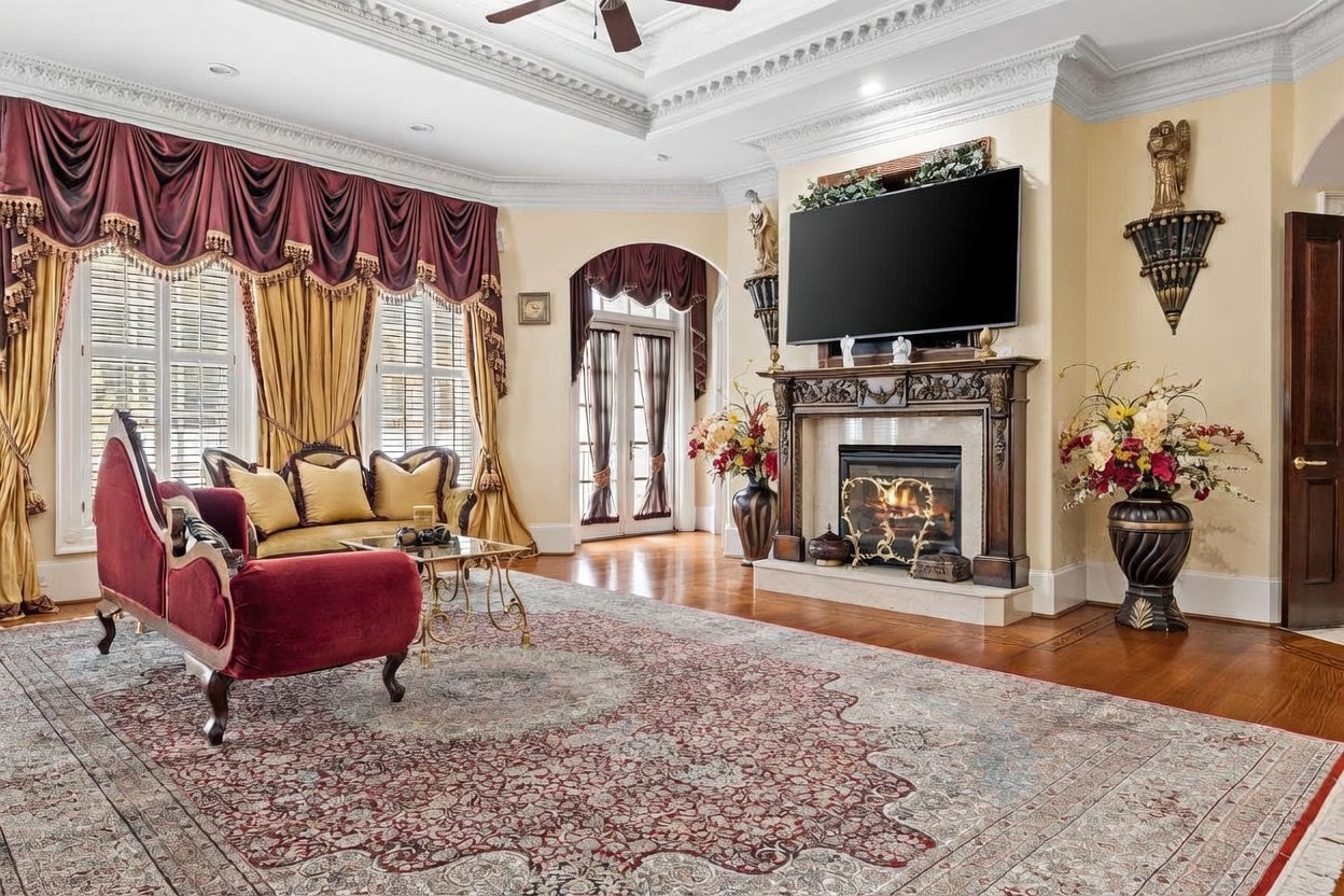 This formal living room features an ornate, traditional aesthetic with a large, intricate area rug centered on hardwood floors. A prominent fireplace with a detailed wood mantel serves as the focal point, topped by a large television and flanked by decorative wall sconces. The space is furnished with a velvet-upholstered chaise lounge and a sofa, all framed by heavy, draped window treatments and crown molding.