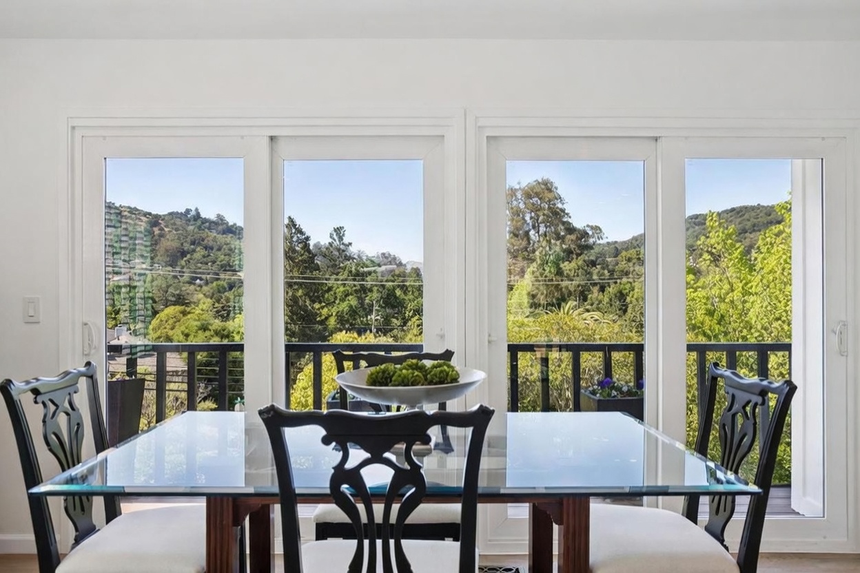 This dining room features a glass-topped table surrounded by dark-framed chairs with light-colored upholstery. The space is defined by a wall of large windows that provide an expansive, unobstructed view of the lush green hills and trees outside. The bright, natural light creates an airy and inviting atmosphere, highlighting the clean lines of the furniture and the serene outdoor scenery.