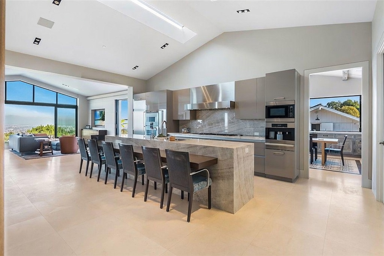 This modern, open-concept kitchen features a large central island with a stone countertop and seating for six, serving as the focal point of the room. The space is characterized by sleek, high-gloss cabinetry, stainless steel appliances, and a vaulted ceiling with recessed lighting. A wide opening leads to an adjacent living area with large windows that provide an expansive view of the outdoors.