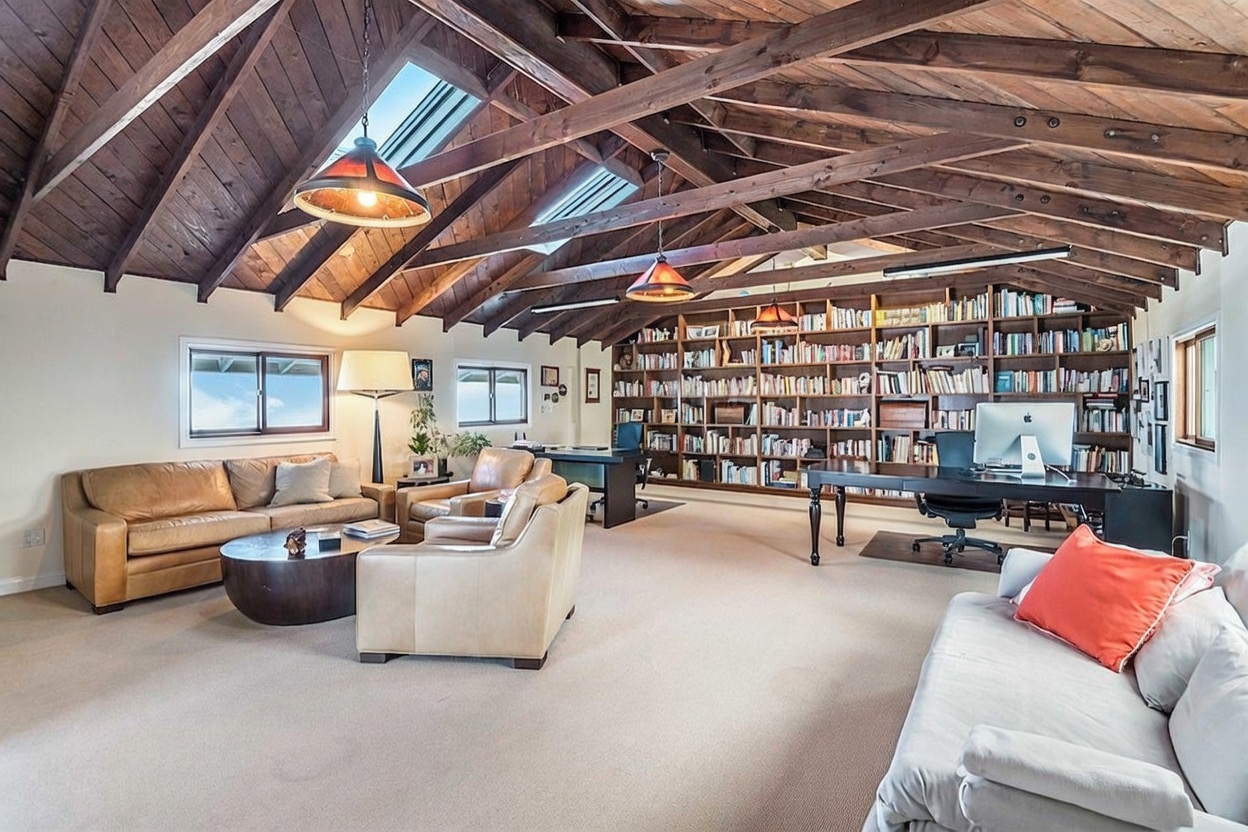 This expansive, multi-functional room features a dramatic vaulted ceiling with exposed dark wood beams and integrated skylights. The space is furnished with a comfortable leather sofa seating area, two desks, and an extensive wall-to-wall bookshelf, creating a sophisticated home office and library environment. The room is bathed in natural light from multiple windows and skylights, highlighting the warm, inviting atmosphere of this versatile living space.