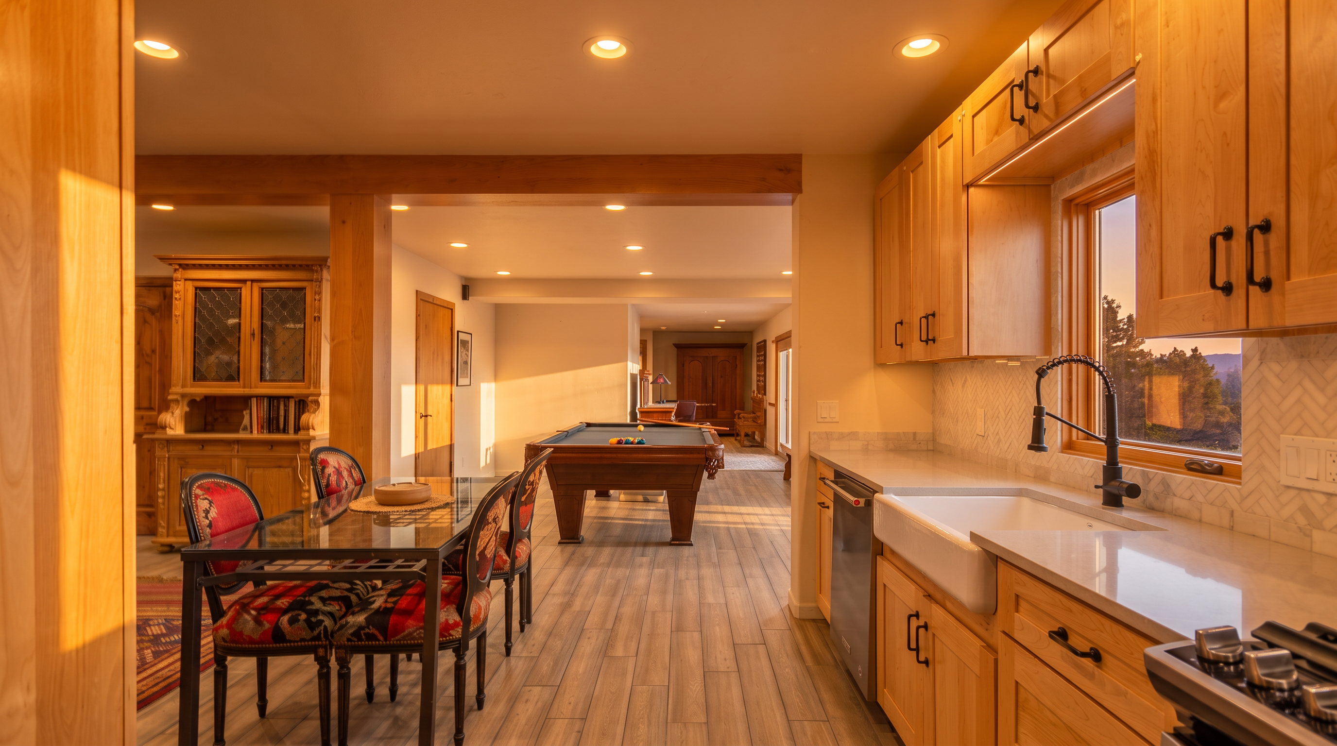 This spacious kitchen area flows into an open-concept living space that includes a formal dining set and a billiards room. The kitchen itself features natural wood cabinetry, a white farmhouse sink, and a stylish herringbone-patterned backsplash, complemented by modern black matte hardware. The warm lighting and hardwood flooring create a welcoming, functional atmosphere that blends traditional elements with a social, entertaining-focused layout.