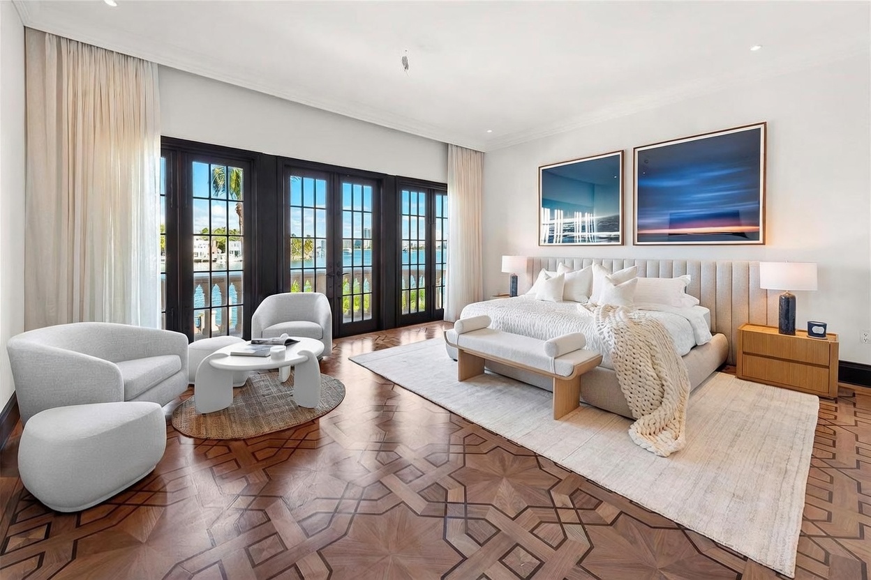 This spacious primary bedroom features intricate patterned wood flooring and a large, upholstered headboard centered between two matching nightstands with modern lamps. A seating area with two curved armchairs and a low coffee table sits near large, dark-framed glass doors that open to a balcony with water views. The room is decorated in a neutral palette with soft textures, including a chunky knit throw on the bed and two large, abstract blue-toned artworks on the wall.
