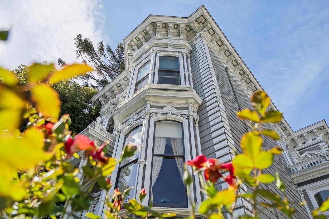 This low-angle, upward-looking shot captures the ornate Victorian architecture of a historic home, emphasizing its intricate trim and bay window structure. The facade features light-colored horizontal siding and elaborate decorative woodwork, framed by soft-focus, vibrant green and red foliage in the foreground. The perspective highlights the building's verticality against a bright blue sky, creating a classic and elegant impression.