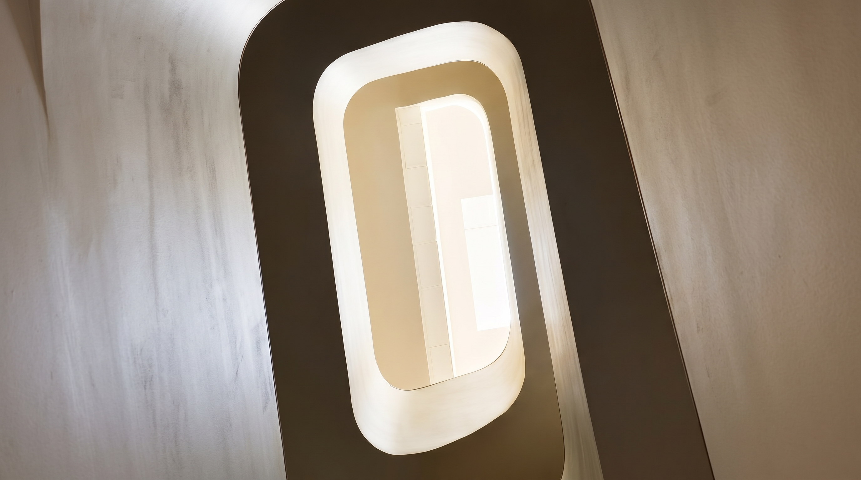 This image captures a striking, sculptural staircase from a high-angle, top-down perspective, emphasizing its fluid, organic curves. The minimalist design features smooth, light-toned surfaces that create a sense of modern elegance and architectural sophistication. The central void draws the eye downward, highlighting the seamless integration of form and function in this contemporary interior space.
