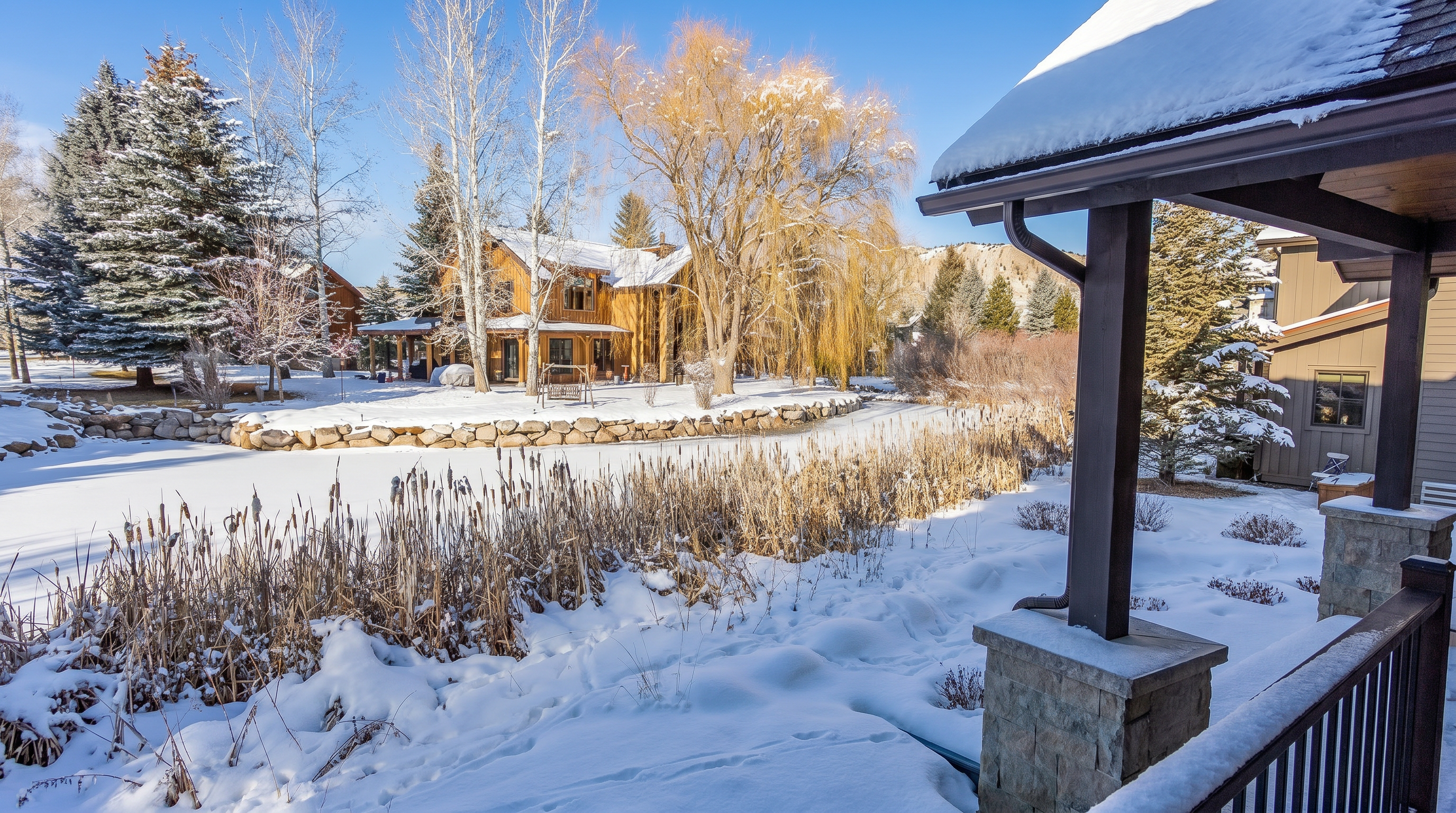 This scenic rear view captures a tranquil winter landscape featuring a snow-covered yard and a frozen pond bordered by tall, dried cattails. In the background, a rustic wooden home stands nestled among bare trees, with a dramatic, sunlit mountain ridge rising against a clear blue sky. The perspective is framed from a covered porch, emphasizing the peaceful, secluded atmosphere of the property.