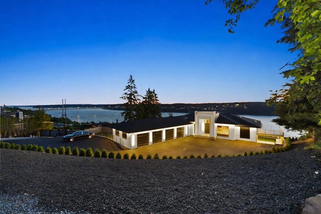 This modern, single-story residence is captured at dusk, showcasing a sleek architectural design with a multi-car garage and a prominent, illuminated entryway. The home is situated on a hillside overlooking a tranquil body of water and a distant bridge, with a dark gravel foreground leading up to the paved driveway. The composition emphasizes the home's striking silhouette against the deep blue twilight sky, highlighting its contemporary aesthetic and prime waterfront location.