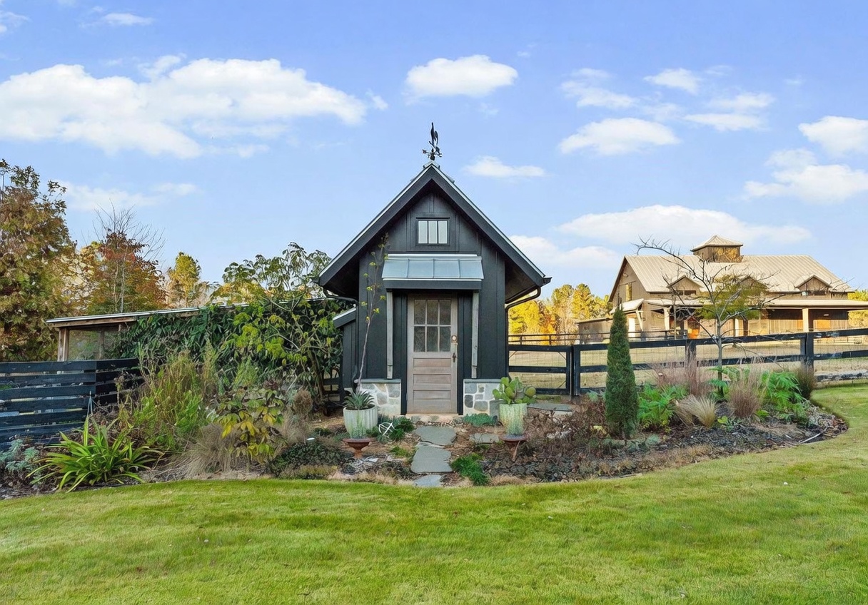 This image captures a charming, dark-painted garden shed or outbuilding featuring a steep gable roof, a weather vane, and a stone-accented entryway. The structure is nestled within a lush, landscaped garden bed filled with various shrubs, grasses, and potted plants, with a stone path leading to the door. In the background, a larger barn-style building and a black wooden fence complete the rustic, pastoral aesthetic of the property.
