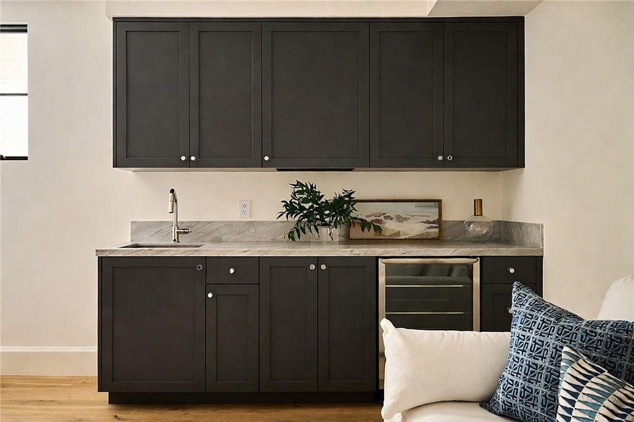 This image showcases a sleek, modern wet bar area featuring dark charcoal cabinetry paired with a light-toned stone countertop and backsplash. The space includes a built-in beverage refrigerator, a stainless steel sink with a modern faucet, and decorative greenery, creating a sophisticated and functional entertaining nook. The perspective is a straight-on, eye-level shot that highlights the clean lines and contemporary aesthetic of the cabinetry and finishes.