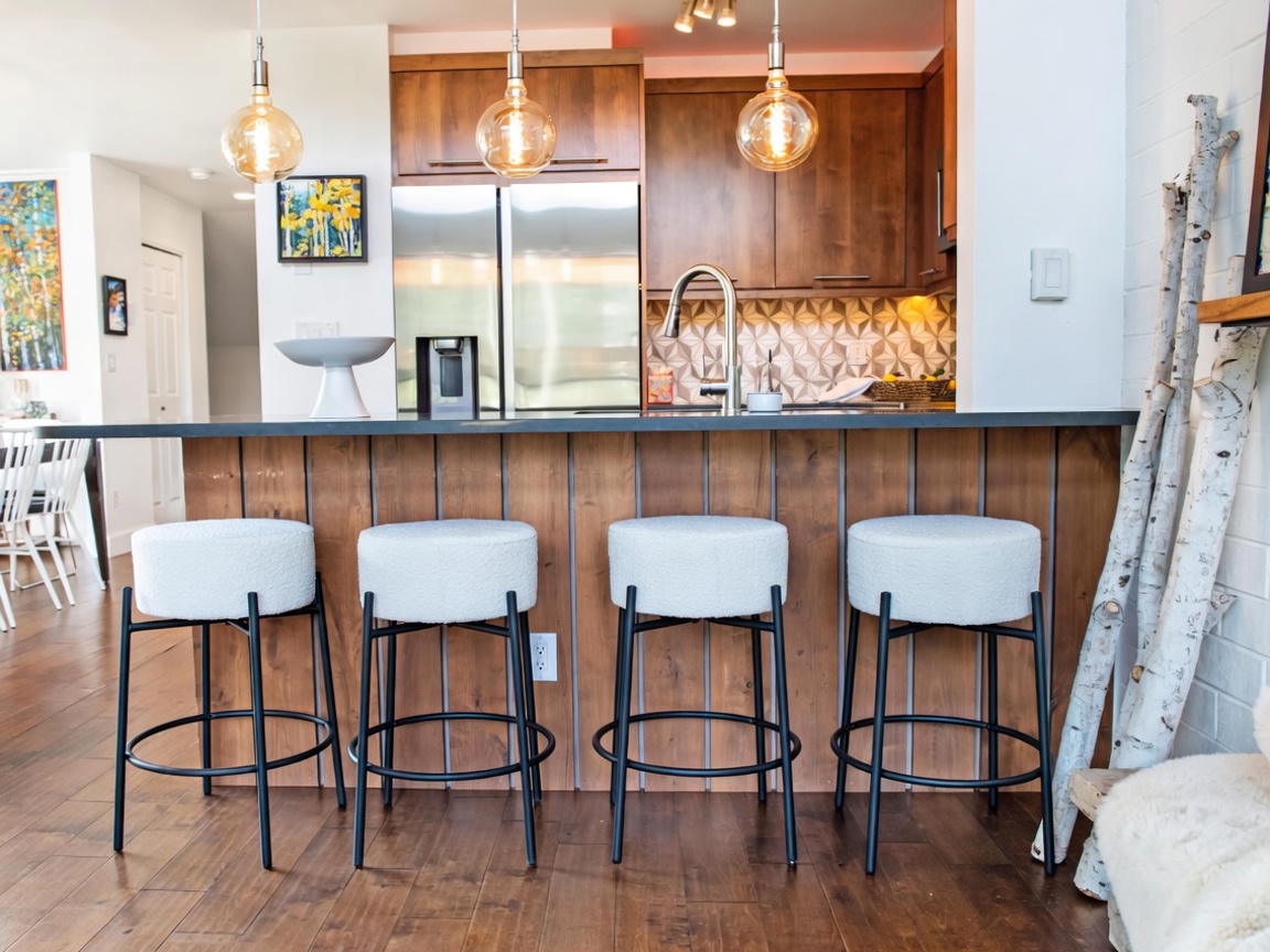 This modern kitchen features a breakfast bar with four white, textured stools set against a wood-paneled base. The space is illuminated by three hanging pendant lights with exposed bulbs, highlighting the stainless steel refrigerator and patterned tile backsplash. To the right, a decorative arrangement of birch logs adds a rustic, organic touch to the contemporary design.