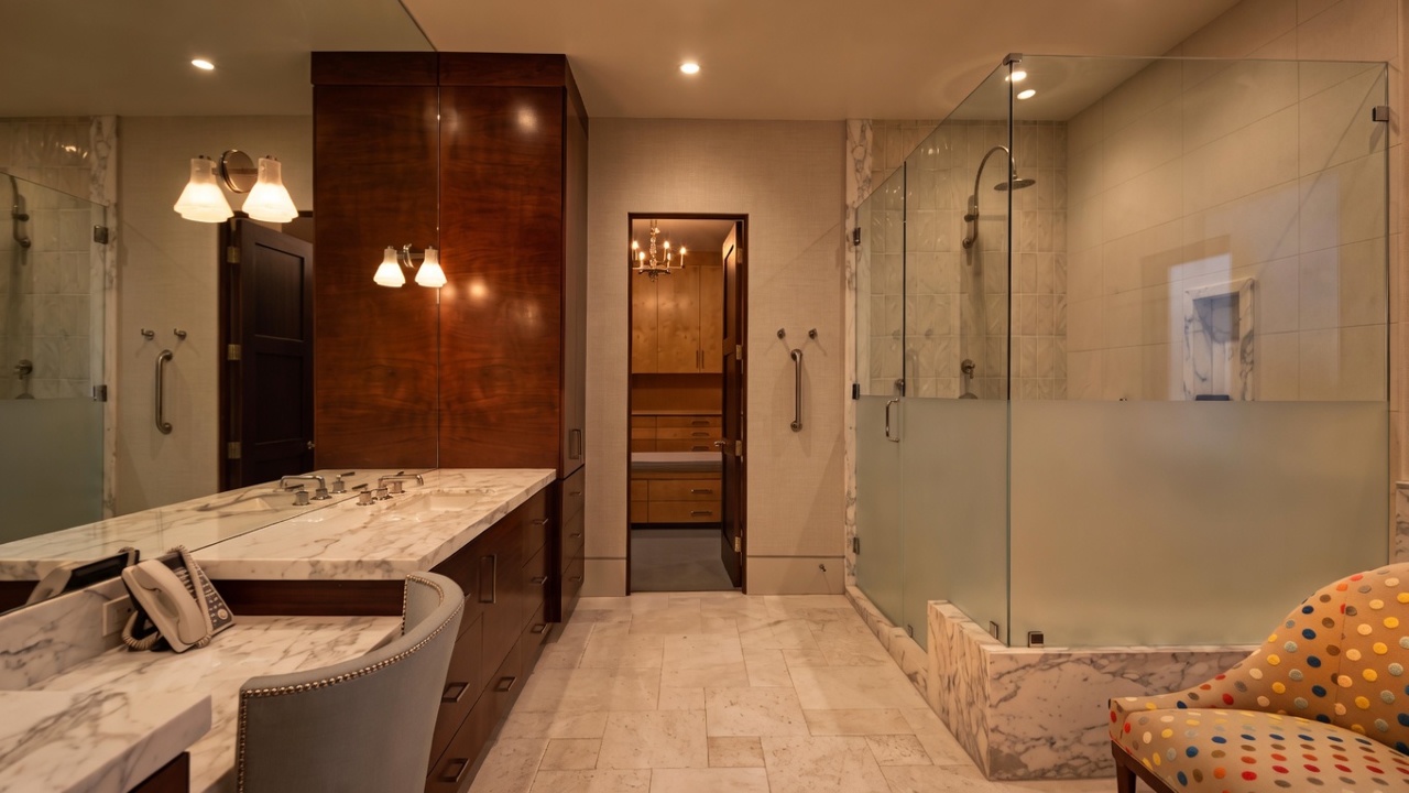This spacious primary bathroom features a long vanity with a stone countertop, a large mirror, and a dedicated makeup area with a chair. To the right, a glass-enclosed shower with a frosted lower section provides privacy while maintaining an open feel. The room is finished with light-toned stone flooring and leads into a walk-in closet, creating a luxurious and functional layout.