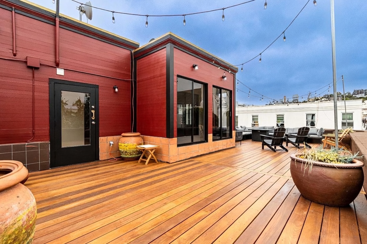 This expansive rooftop deck features warm-toned wood decking and a striking red exterior wall with black trim. The space is furnished with outdoor seating, large decorative planters, and string lights overhead, creating an inviting atmosphere for entertaining. The perspective captures the deck from a low angle, highlighting the clean lines of the architecture and the open-air layout.