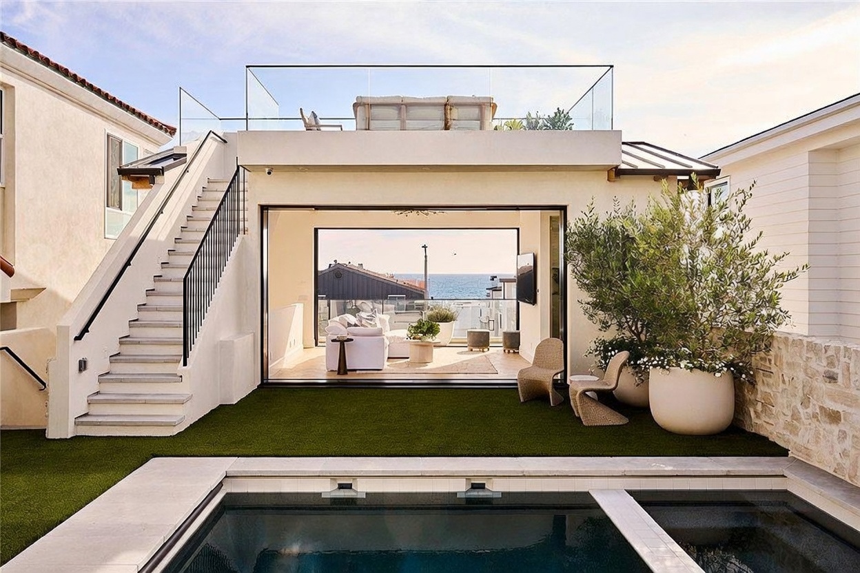 This contemporary outdoor living space features a sleek swimming pool in the foreground leading to a manicured artificial turf lawn. A modern structure with floor-to-ceiling glass walls opens to reveal a bright interior living area, while an external staircase provides access to a rooftop deck enclosed by glass railings. The scene is framed by a large potted olive tree and a stone-clad wall, creating a sophisticated and inviting coastal atmosphere.