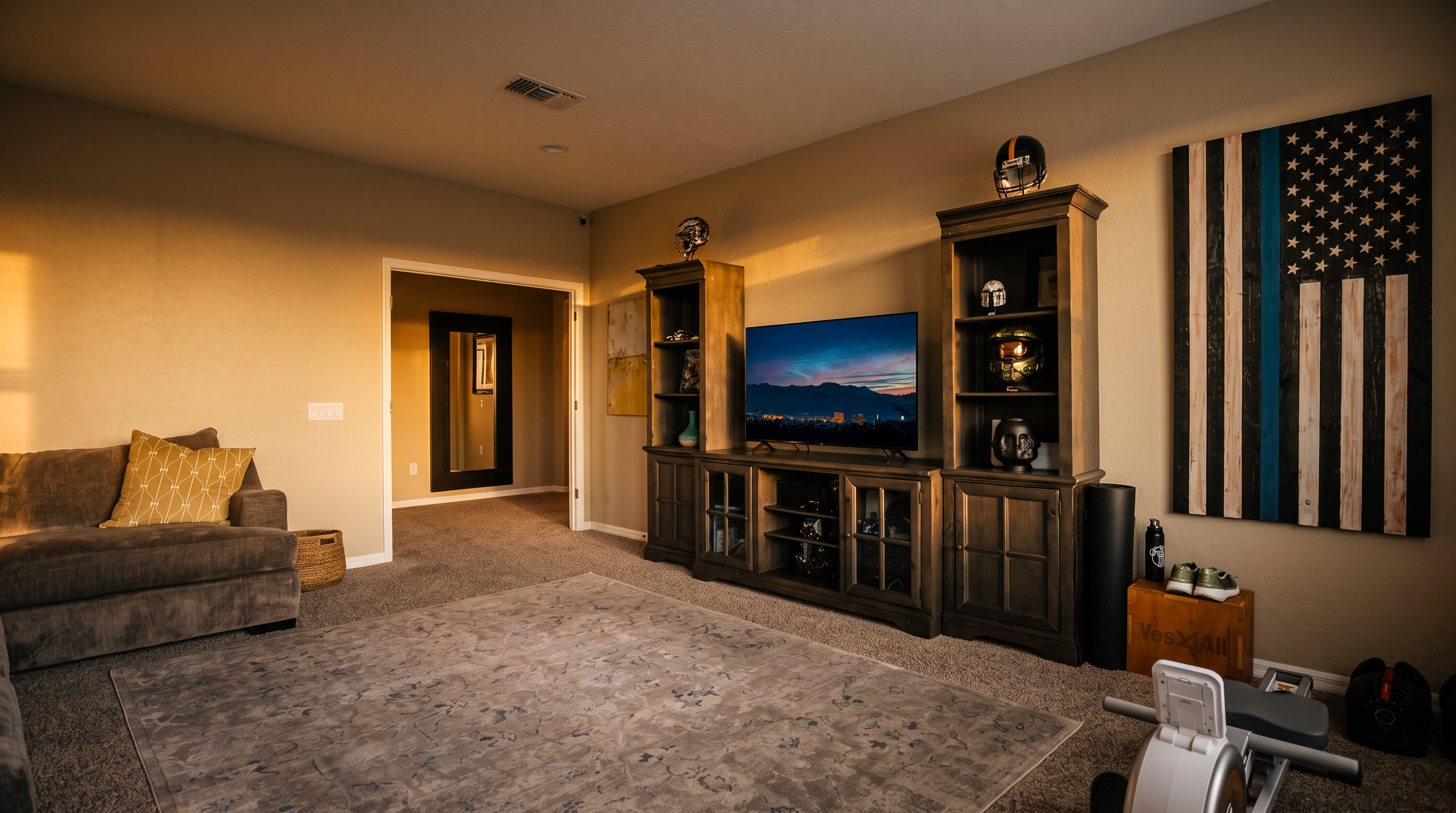 This versatile living space features a dark wood entertainment center housing a television, flanked by shelving units displaying various collectibles and sports memorabilia. A comfortable grey sofa with a patterned throw pillow sits on a large, neutral-toned area rug, while a workout machine and a wooden crate are positioned to the side. The room is decorated with a large, rustic-style American flag wall art piece and is illuminated by recessed ceiling lights, creating a functional multi-purpose area.